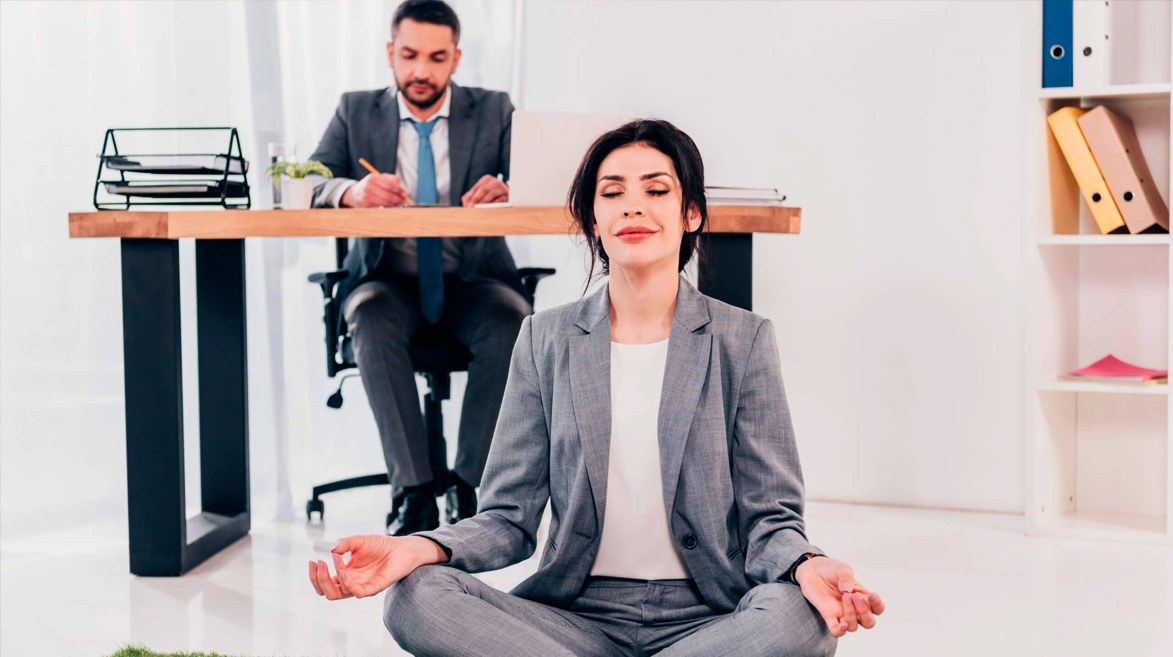 Foto em um escritório; em segundo plano há um homem sentado em frente a uma mesa e sobre a mesa um notebook; e em primeiro plano uma mulher sentada no chão em posição de meditação e com os olhos fechados e um sorriso discreto;