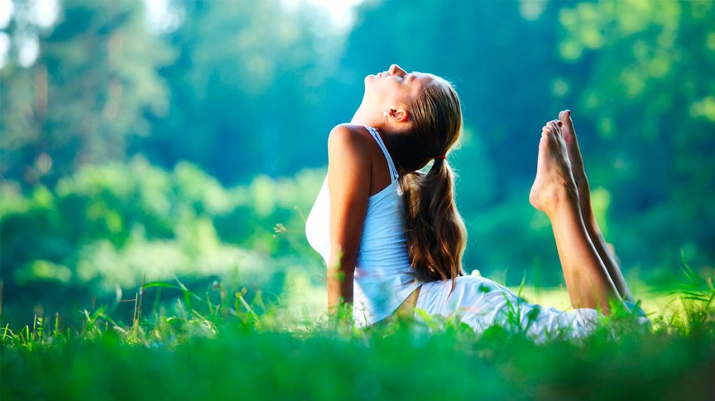 Mulher num parque alongando-se sobre a grama num dia de sol. Ela está deitada de barriga para baixo, com os pés e cabeça em direção ao céu, alongando tronco, tórax e pés.