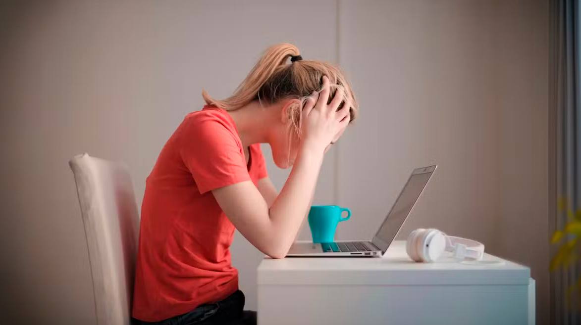 Mulher sentada em frente a uma pequena mesa de escritório e com os cotovelos apoiados a mesa, as mãs sobre a cabeça, rosto olhando para baixo, sobre a mesa, há uma caneca, um notebook com a tela aberta e um fone de ouvido. A foto representa uma pessoa com burnout.