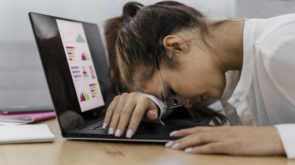 A imagem mostra uma mulher com aparência cansada, debruçada sobre um laptop. Ela está usando óculos, que estão levemente caídos no rosto. A tela do laptop exibe gráficos coloridos, indicando possivelmente um ambiente de trabalho ou estudo. A mulher parece estar exausta, com a cabeça apoiada no teclado e uma das mãos sobre o laptop.