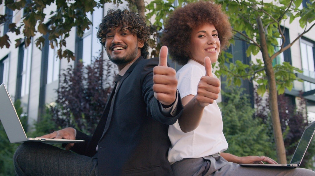 Na imagem, vemos duas pessoas sentadas de costas uma para a outra, ao ar livre, em um ambiente corporativo cercado por árvores. À esquerda, um homem com cabelo cacheado, barba curta e pele morena, veste um terno preto e sorri enquanto levanta o polegar direito, expressando aprovação. À direita, uma mulher com cabelo volumoso e crespo, pele clara e vestida com uma camisa branca, também sorri e levanta o polegar direito. Ambos estão utilizando laptops, simbolizando uma atmosfera de trabalho e cooperação. A expressão de ambos é positiva e transmite uma mensagem de confiança e sucesso. A imagem reflete um ambiente de trabalho saudável e colaborativo, ideal para ilustrar a temática de empresas comprometidas com a saúde mental.
