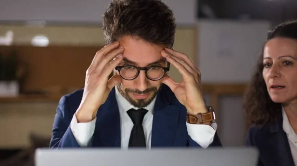 Na imagem, um homem de cabelos curtos e barba, usando óculos de armação escura e vestindo um terno azul marinho, está sentado em frente a um laptop, com uma expressão visivelmente preocupada. Ele está com as mãos na cabeça, pressionando as têmporas, como se estivesse lidando com uma situação estressante ou difícil. Ao lado dele, uma mulher de cabelos castanhos, aparentando ser uma colega de trabalho, observa a situação com uma expressão séria e preocupada. O ambiente parece ser um escritório, com iluminação suave e um fundo desfocado, sugerindo um local de trabalho moderno. Esta imagem ilustra o impacto do estresse no ambiente de trabalho e reforça a importância de empresas promoverem a saúde mental de seus colaboradores. Este conteúdo está associado ao tema "Empresas comprometidas com a saúde mental receberão certificado especial do governo federal", destacando a relevância de iniciativas que priorizem o bem-estar emocional no ambiente corporativo.