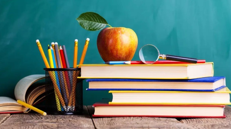 A imagem mostra uma mesa de madeira com materiais escolares organizados. No lado esquerdo, há um suporte para lápis cheio de canetas, lápis e um marcador amarelo. Ao centro, estão três livros empilhados horizontalmente, e sobre o livro superior há uma maçã vermelha com uma folha verde. Ao lado da maçã, encontra-se uma lupa posicionada sobre os livros. O fundo é uma superfície verde, lembrando um quadro escolar. Essa imagem remete ao ambiente acadêmico e simboliza o conhecimento e a busca por aprendizado, temas que podem ser associados à importância de incluir a saúde mental como pauta nas empresas, considerando o bem-estar e a educação sobre o tema no ambiente de trabalho.