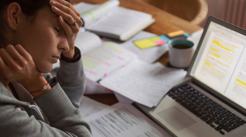 A imagem mostra uma mulher jovem, de pele clara e cabelos castanhos presos em um rabo de cavalo, com expressão de cansaço e preocupação. Ela está sentada à mesa, cercada por vários papéis espalhados e livros abertos. Com a mão esquerda, ela cobre parte do rosto, pressionando a testa, enquanto mantém os olhos fechados, sugerindo exaustão ou sobrecarga mental. Em frente a ela, há um laptop aberto com texto destacado em amarelo na tela, indicando que ela está estudando ou trabalhando. Também há uma xícara de café à direita do laptop, reforçando a ideia de que ela está tentando manter-se acordada e concentrada.