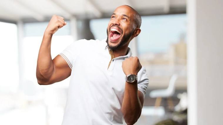 A imagem mostra um homem com expressão de alegria e comemoração. Ele veste uma camisa polo branca e levanta os braços com os punhos cerrados, expressando entusiasmo e vitória. Seu rosto exibe um grande sorriso e os olhos estão direcionados para cima, reforçando a sensação de conquista. O ambiente ao fundo é claro e desfocado, sugerindo que ele está em um espaço moderno e bem iluminado, possivelmente um escritório. Essa imagem transmite sentimentos de felicidade, motivação e sucesso, sendo ideal para temas relacionados a saúde, bem-estar e realização pessoal em ambientes corporativos.