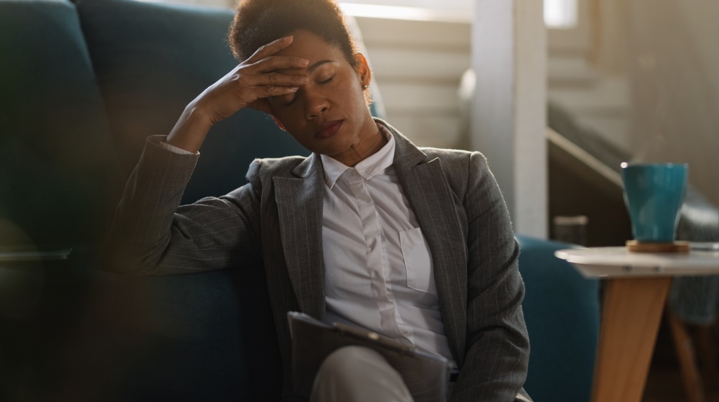 A imagem mostra uma mulher sentada em um ambiente interno, com expressão de cansaço e esgotamento. Ela veste um blazer cinza e camisa branca, sugerindo um contexto profissional. Sua mão esquerda está apoiada na testa, enquanto seus olhos estão fechados, reforçando a ideia de sobrecarga emocional ou física. Ao fundo, há móveis e uma xícara azul sobre uma mesa lateral, criando um ambiente acolhedor, mas contrastando com seu estado emocional. A iluminação é suave e natural, contribuindo para um tom reflexivo e introspectivo.