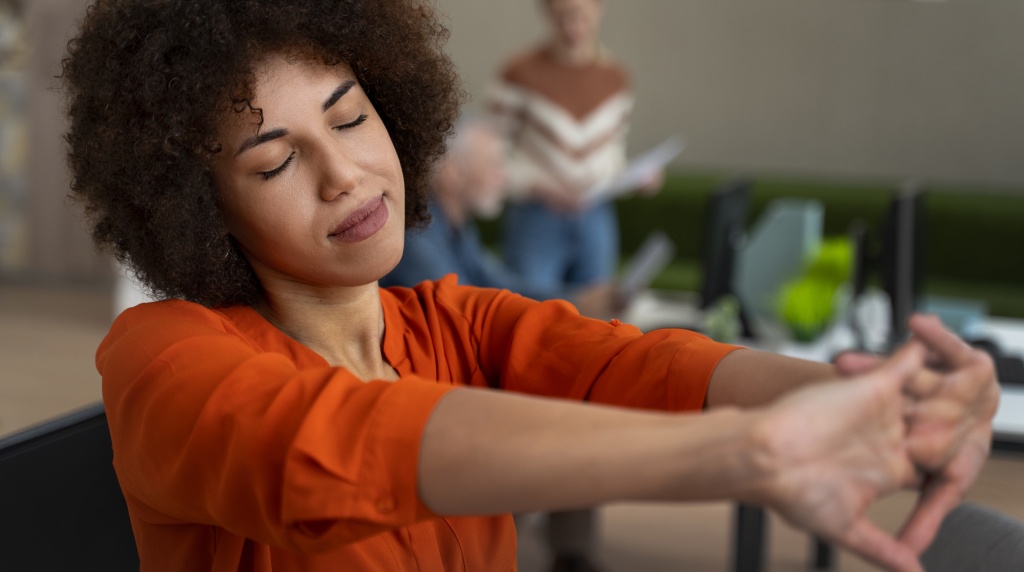 A imagem mostra uma mulher jovem, de cabelos cacheados, sentada em um ambiente corporativo enquanto se alonga com os olhos fechados, transmitindo uma sensação de pausa e cuidado consigo mesma. Ela veste uma camisa laranja vibrante, que contrasta com o cenário do escritório ao fundo, onde outras pessoas trabalham e interagem. O ambiente é moderno e iluminado, sugerindo um local de trabalho que valoriza o bem-estar. O ato de alongar as mãos simboliza a importância de pequenos momentos de autocuidado para aliviar o estresse no dia a dia corporativo.