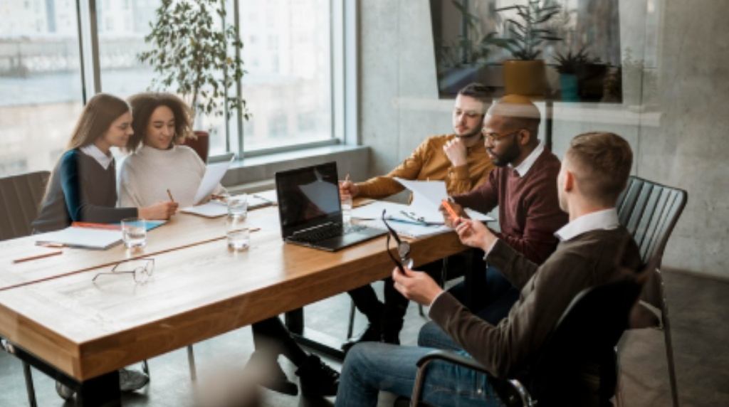 Cinco pessoas estão reunidas em uma mesa de madeira em um ambiente corporativo moderno, com uma grande janela ao fundo que permite a entrada de luz natural. O grupo é diverso e composto por homens e mulheres de diferentes etnias, todos engajados em uma conversa. Duas mulheres à esquerda analisam documentos juntas, enquanto três homens à direita discutem segurando papeis e óculos. No centro da mesa, há um notebook aberto, copos com água e materiais de anotação, sugerindo um ambiente colaborativo e produtivo. O espaço tem uma decoração sofisticada, com plantas e elementos contemporâneos. A cena transmite um clima de diálogo, troca de ideias e planejamento estratégico. A imagem ilustra a importância do olhar organizacional para a saúde mental no ambiente de trabalho. O engajamento e a troca de ideias refletem um ambiente onde o bem-estar dos colaboradores é valorizado, promovendo um espaço mais saudável e produtivo.