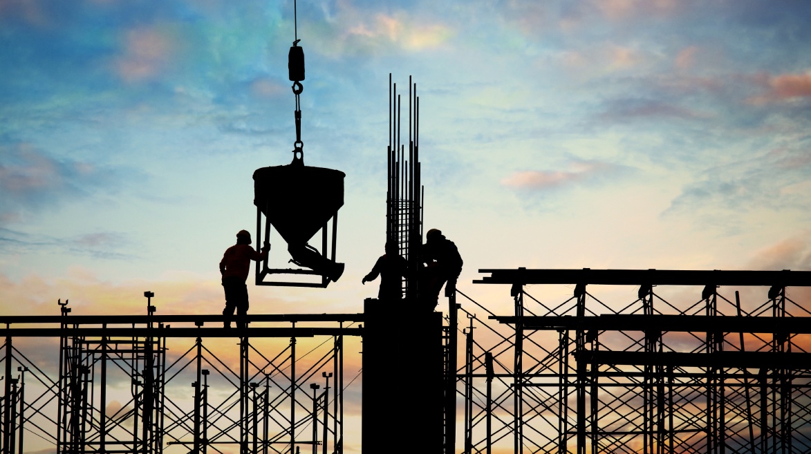 A imagem mostra a silhueta de três trabalhadores em uma obra civil durante o pôr do sol, em cima de uma estrutura de andaimes metálicos e pilares em construção. Eles operam um guindaste que ergue um recipiente provavelmente contendo concreto. O céu ao fundo apresenta tons suaves de azul, amarelo e laranja. Essa cena reforça visualmente a importância da Avaliação Psicossocial na construção civil, destacando a necessidade de atenção à segurança e saúde mental dos trabalhadores expostos a condições de risco em altura e operações complexas.