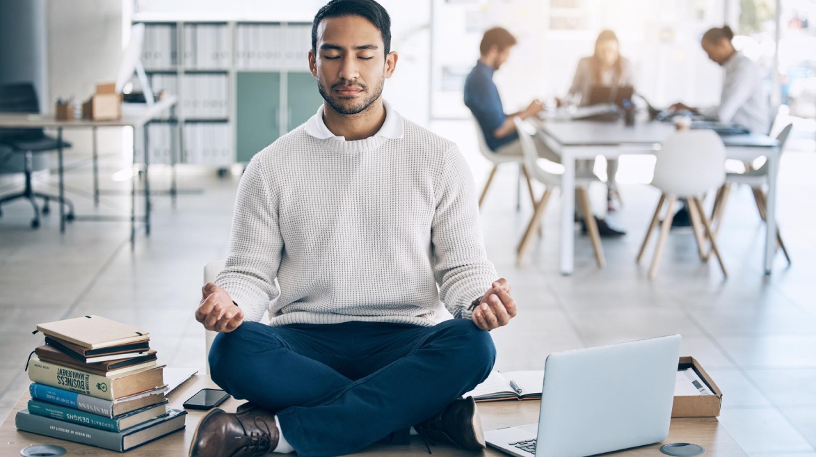 A imagem retrata um jovem universitário sentado de pernas cruzadas sobre uma mesa em um ambiente de estudo ou trabalho. Ele veste um suéter branco e calça azul, mantendo os olhos fechados e as mãos apoiadas nos joelhos em um gesto meditativo. Ao redor dele, há pilhas de livros, um laptop aberto e um smartphone, sugerindo um contexto acadêmico ou profissional. No fundo, outros estudantes ou colegas trabalham em grupo, desfocados, contrastando com a serenidade do protagonista. A luz natural entra no ambiente, reforçando a sensação de tranquilidade e foco. A imagem simboliza a prática do mindfulness como ferramenta para a regulação emocional e o alívio do estresse em meio às pressões da vida acadêmica.