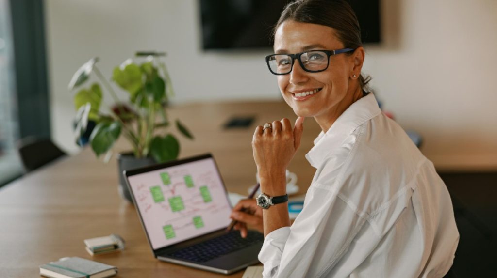 Uma mulher profissional sorri de maneira confiante enquanto trabalha em um ambiente de escritório moderno e bem iluminado. Ela usa óculos de grau e veste uma camisa social branca, transmitindo uma imagem de profissionalismo e organização. Sentada à mesa, ela interage com um laptop, onde uma tela exibe um diagrama ou fluxograma, indicando planejamento estratégico ou organização de tarefas. Ao lado do computador, há cadernos e uma planta verde, elementos que remetem a um ambiente de trabalho equilibrado e produtivo. A composição da imagem sugere boas práticas para manter a produtividade no trabalho, incluindo organização, planejamento visual e um ambiente agradável com toques naturais. O sorriso e a postura relaxada reforçam a ideia de satisfação e eficiência na rotina profissional.