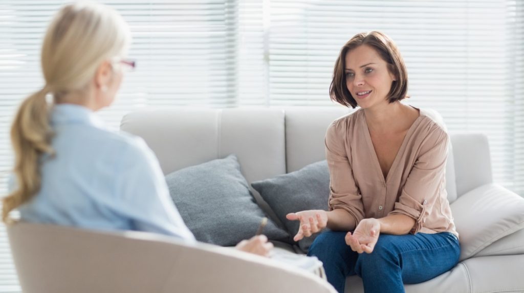 Na imagem, duas mulheres estão sentadas em um sofá claro, num ambiente bem iluminado e tranquilo. A mulher de cabelos escuros, usando blusa rosada e jeans, fala de forma descontraída enquanto faz gestos suaves com as mãos. A outra, loira, está de costas para a câmera, inclinada para frente em atitude de escuta. A cena transmite acolhimento, confiança e foco no bem-estar emocional, refletindo a importância de ações de saúde mental direcionadas às mulheres em ambientes corporativos.