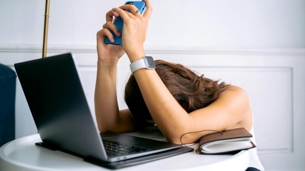 Mulher sentada à mesa, com a cabeça apoiada nos braços em sinal de exaustão. Ela segura um celular azul acima da cabeça, enquanto à sua frente há um notebook aberto e um caderno fechado. A expressão corporal transmite cansaço extremo e possível sobrecarga emocional, representando o esgotamento mental. O ambiente é claro e minimalista, reforçando a sensação de isolamento. O cenário ilustra o tema do esgotamento no trabalho e seu impacto na saúde mental das mulheres.