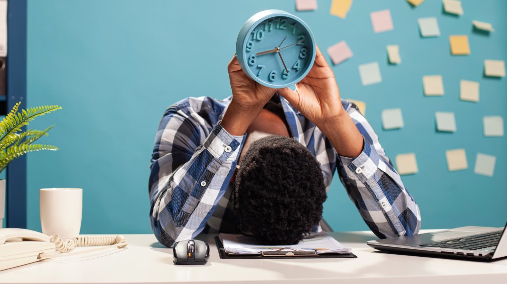 Na imagem, uma pessoa sentada à mesa de trabalho apoia a cabeça sobre os braços, segurando acima dela um relógio azul que marca pouco antes das 8 horas. A cena transmite exaustão e sobrecarga, com um ambiente de escritório ao fundo, incluindo laptop, telefone, mouse, planta decorativa e uma parede azul repleta de post-its coloridos, sugerindo múltiplas tarefas pendentes. A expressão corporal reforça a ideia de cansaço extremo e pressão pelo tempo, associada ao tema do burnout como potencial doença ocupacional da segunda metade do século XXI.